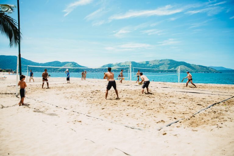 Pessoas jogando beach tennis em uma praia ensolarada no Brasil, com o mar e as montanhas ao fundo, representando o estilo de vida ativo e tropical do esporte.